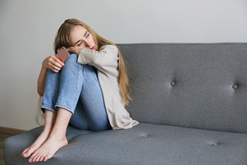 Portrait of beautiful young woman with depressed facial expression sitting on grey textile couch...