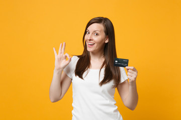 Portrait of pretty young woman in white casual clothes showing OK gesture, holding credit bank card isolated on yellow orange wall background in studio. People lifestyle concept. Mock up copy space.