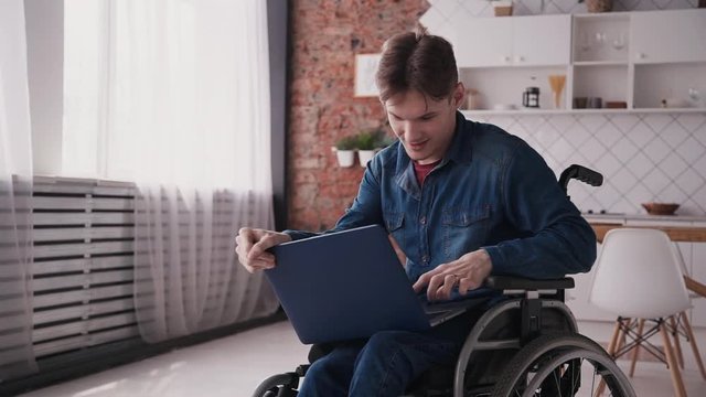 Young or adult disabled man sitting in comfort wheel chair and spending free time at modern flat with bright and light room inside. Male with calm face holding laptop on knees and working
