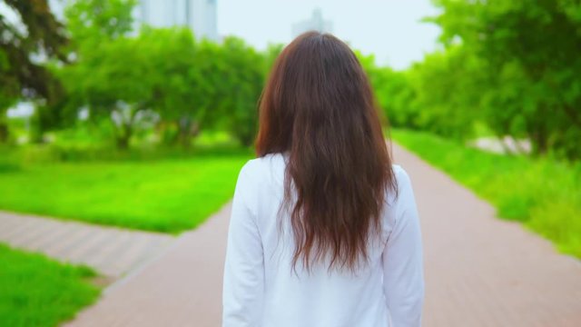 back view woman with long hair walking in city with green trees and modern buildings on the background