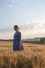 young cute girl in blue dress with collected hair looks at camera in field of ears at sunrise. Foggy dawn in background