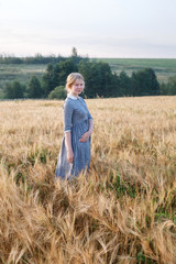 Naklejka na ściany i meble nice happy young girl in blue old-fashioned outdoor dress in golden field of ears at dawn. Green boscage in background.