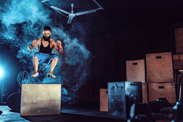 Fit tattoed bearded man jumping onto a box as part of exercise routine. Man doing box jump in the gym. Athlete is performing box jumps.