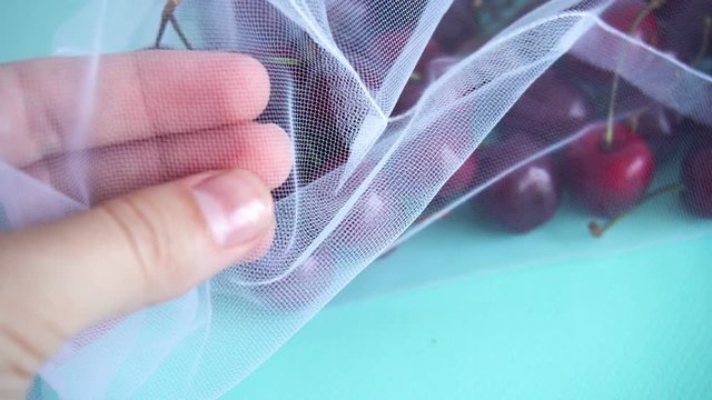 female hand touches cherry in a reusable fabric transparent bag on a blue background in the fight against plastic. close-up texture