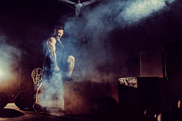 Muscular attractive caucasian bearded tasttoed man lifting kettlebell in a gym. Weight plates, dumbbell and tires in smoke background.
