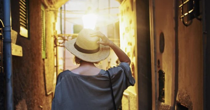 Back view of pretty woman holding hat walking in sunlight in beauty tight old street. Female wanderer traveling around medieval coastal italian town in summer.