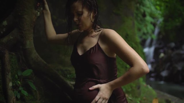 Attractive pregnant woman feels labor contractions pain and concentrates on her breathing after swimming in the river in the tropical jungle. Slow motion.
