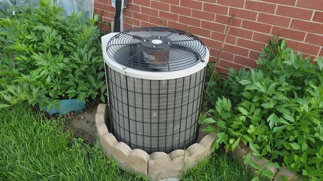 Single Family Home Old Rusty AC Unit Or Air Conditioner External Heat Exchange Unit Spinning And Blowing Air Against Some Plants And Brick Wall.