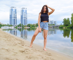 Young brunette girl in denim shorts stands near the river on the beach.