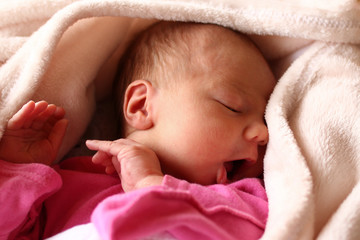 Cute newborn baby in pink baud with an open mouth on her bed under beige blanket.