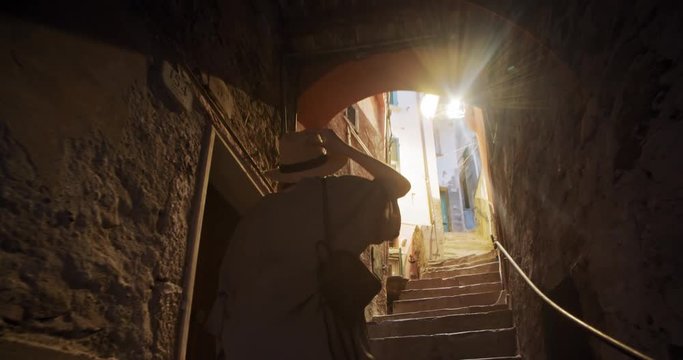 Back view cheerful girl in trendy outfit walking up stone stairs in the beautiful narrow street to the sunlight. Exploring Tuscany, Riomaggiore in summer.
