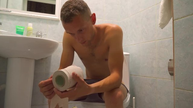 Naked man sitting on the toilet, pensive. Twists in the hands of toilet paper.