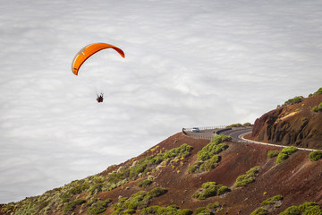 Fototapeta na wymiar a paraglider on a red with yellow stripes paraglider flies in sky with white clouds, Tenerife, Teide