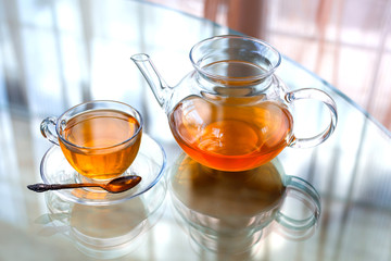 Transparent glass tea cup and pot with spoon on glass table in strong backlit