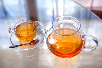 Transparent glass tea cup and pot with spoon on glass table in strong backlit