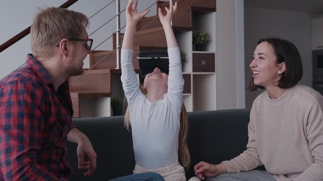 Spending free time at house together. Happy family of three people sitting on sofa or couch inside flat with modern interior room. Little girl using 3d vr helmet with glasses and having fun