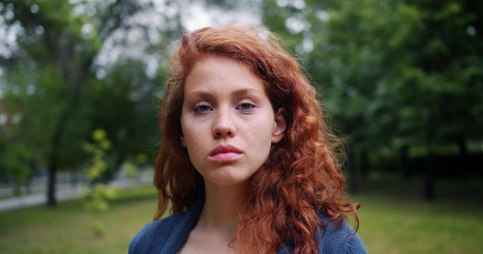 Slow motion portrait of pretty lady with red hair wearing casual clothing standing in the park with serious face and looking at camera. Nature and youth concept.