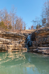 waterfall over green water pond with bare trees and blue sky background