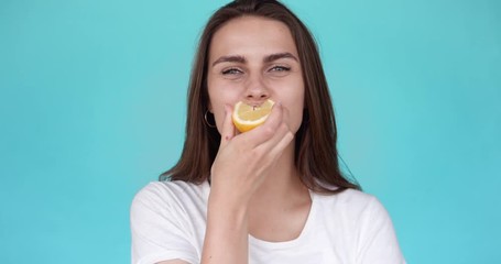 Beautiful girl tries sour lemon. Portrait of a young woman. The girl grimaces while eating a yellow lemon. Slow motion. Turquoise background.