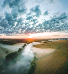  Aerial view over a misty morning over countryside. Fog covering the crop fields and river. Colorful sunrise over the fields and forest. 