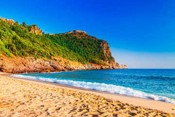 Fototapeta na wymiar Cleopatra beach on sea coast with green rocks in Alanya peninsula, Antalya district, Turkey. Beautiful sunny landscape for tourism with clear water and sand. Alanya Castle on the cliff
