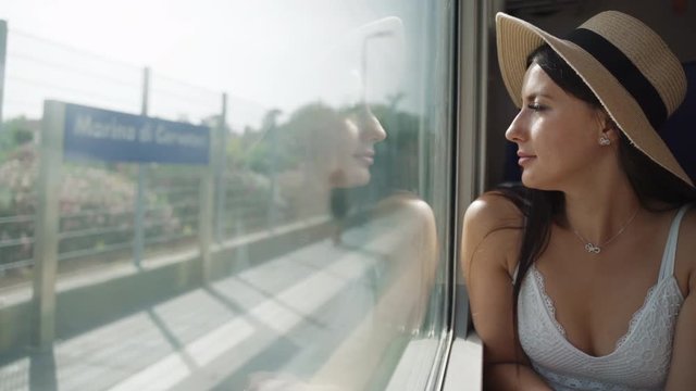Attractive happy girl sitting near the window in a moving train and watching people walking on the train station. Fast vehicle passing railway station and reaching travel destination. Fashionable