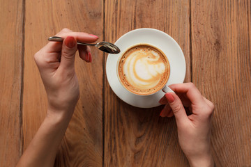 Close-up female hands holding cup with coffee cappuccino with foam with nice pattern. Perfect red gel polish manicure. Wood natural vintage table. Creative color warm post processing