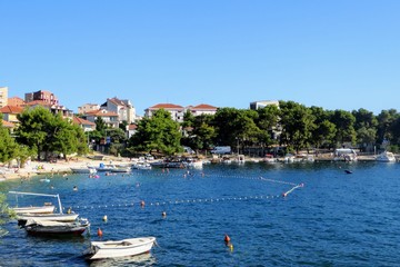Fototapeta na wymiar A beautiful amazing view of a beach on Otok Ciovo or Ciovo island beside Trogir, Croatia. It is a beautiful sunny day with many people swimming in the water and small boats anchored in the bay.