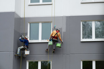 Worker hanging on rope and paints building wall with roller. Painter hanging on cable with paint...