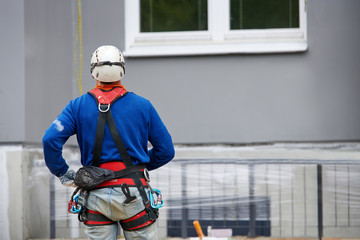 Professional industrial alpinist with rigging equipment, hardhat and safety harness from the back. Painter hanging on cable with paint buckets, industrial climber repairing house facade