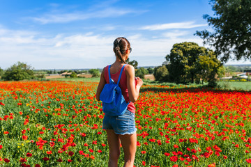 Tourist woman with backpack on the field of red poppies near of Girona, Catalonia, Spain near of Barcelona. Scenic nature landscape in sunny bright day. Famous travel destination in Costa Brava