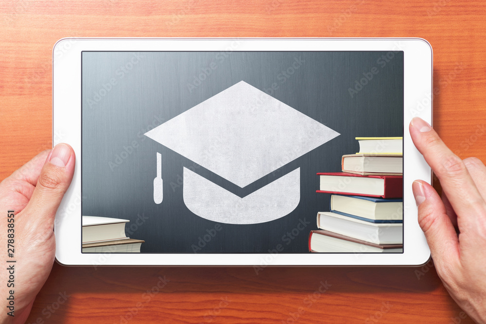 Poster E-learning concept image. Closeup of hands touching white tablet computer on wooden desk. Book, bachelor's cap icon and blackboard. Front view image.