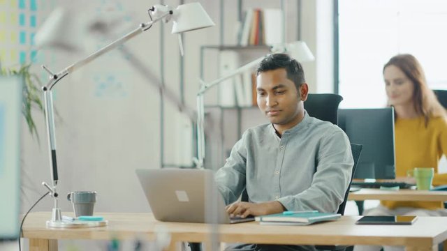Smart and Handsome Indian Information Technology Specialist Sitting at His Desk works on a Laptop. In the Background Modern Office with Diverse Team of Young Professionals Working