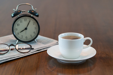 Hot coffee cup and vintage clock newspaper reading glasses on brown wood table