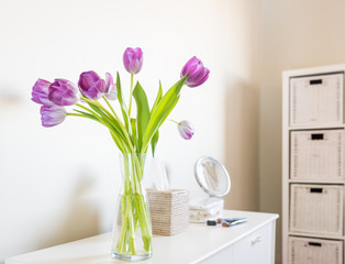 Purple tulips in glass vase on bedroon dressing table with accessories and furniture in background (selective focus)