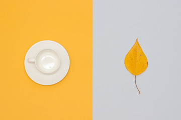 White empty cup with saucer and autumn leaf on yellow and gray background. Copy space Flat lay Mock up Template for your design