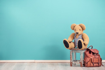 Retro Teddy Bear toy sitting on aged wooden stool and old leather school bag backpack for books front mint blue wall background. Vintage style filtered photo