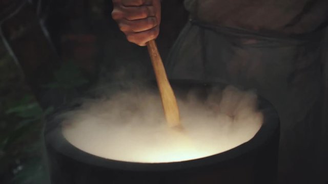 The hand of an old witch is stirring a potion in a bubbling vat with a wooden spoon. Close-up shot with dry ice and steam. Scenery house laboratory for the manufacture of drugs and potions.