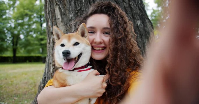 Portrait of attractive lady taking selfie with puppy in park kissing hugging dog looking at camera enjoying summertime. People, photography and animals concept.