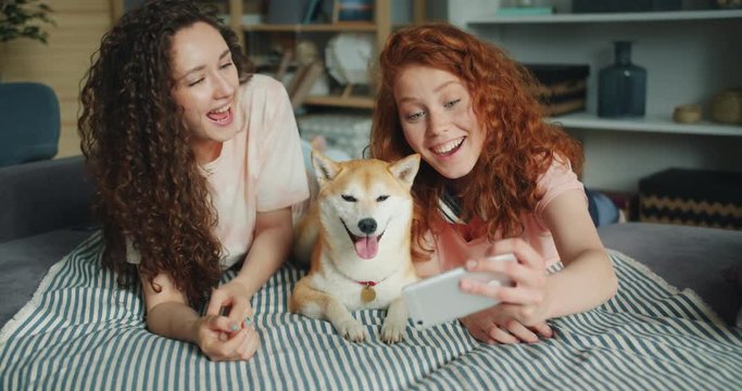 Pretty girls friends are taking selfie with cute dog using smartphone camera lying on couch together having fun. Modern technology, people and photographs concept.