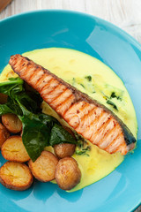 Close up of a grilled salmon steak with baked baby potatoes, spinach and coconut milk gravy served on a bright plate