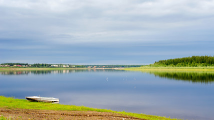 Seagulls fly about a small boat stands on the shore of the Northern Yakut river vilyu near the village of ulus Suntar with houses along the water under a cloudy sky.