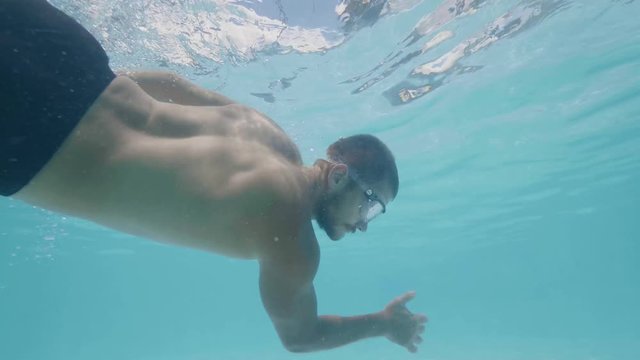 Underwater view young man in goggles swimming in blue water pool. Bearded swimmer swimming in pool slow motion.