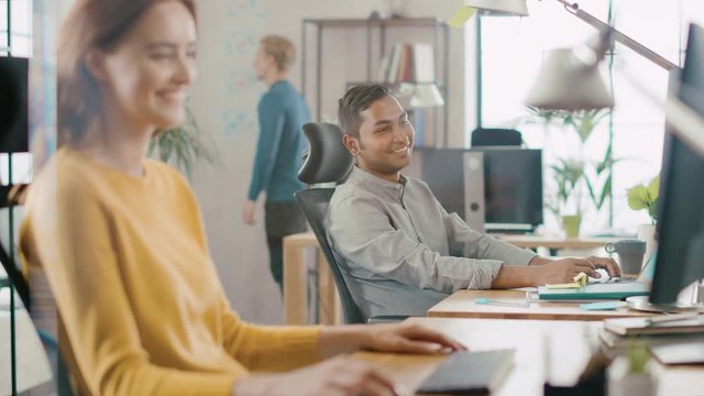 Beautiful and Creative Young Woman Sitting at Her Desk Using Laptop Computer. She Chats, Smiles and Jokes with Her Friendly Colleague. Office where Diverse Team of Young Professionals Work 
