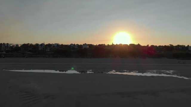 aerial view of a sunrise on the beach of rota, cadiz, you can see 3 people sitting on the sand and another person walking, in the fodo you see houses and trees. The sun is at the bottom