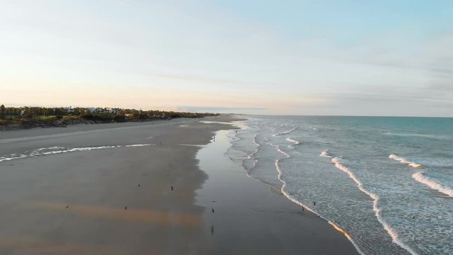 aerial view of a sunrise on the beach of rota, cadiz, you can see 3 people sitting on the sand and another person walking, in the fodo you see houses and trees, waves breaking on the shoregulls flying