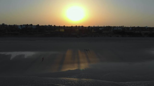 aerial view of a sunrise on the beach of rota, cadiz, you can see 3 people sitting on the sand and another person walking, in the fodo you see houses and trees. The sun is at the bottom