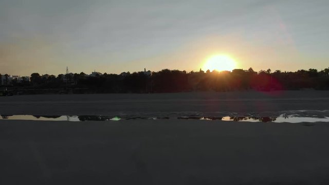 aerial view of a sunrise on the beach of rota, cadiz, you can see 3 people sitting on the sand and another person walking, in the fodo you see houses and trees. The sun is at the bottom