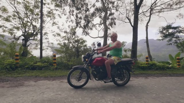 Mature man driving motorcycle on mountain road in asian countryside. Tourist man traveling on motorbike while moto trip on green hills and rainforest landscape.