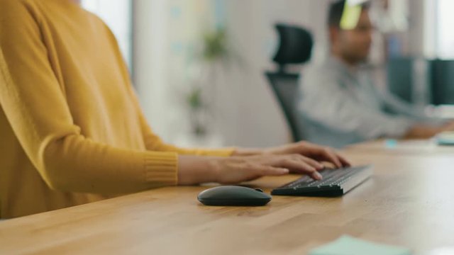Anonymous Young Woman Sitting at Her Desk Using Laptop Computer. Focus on Hands Typing on Keyboard. In the Background Bright Office where Diverse Team of Young Professionals Work 